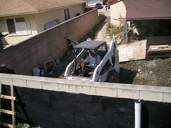White skid-steer loader operating behind a waterproofed retaining wall with drainage pipe at a residential site