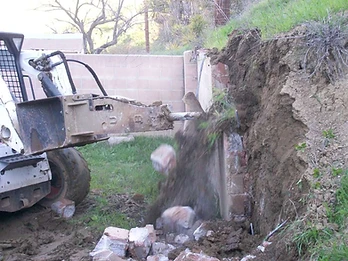 White skid-steer loader with hydraulic hammer demolishing a concrete block retaining wall with dust and debris in mid-air
