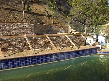 Tiered stone veneer retaining wall with wooden bracing supports, pool with blue solar cover, and dirt hillside behind