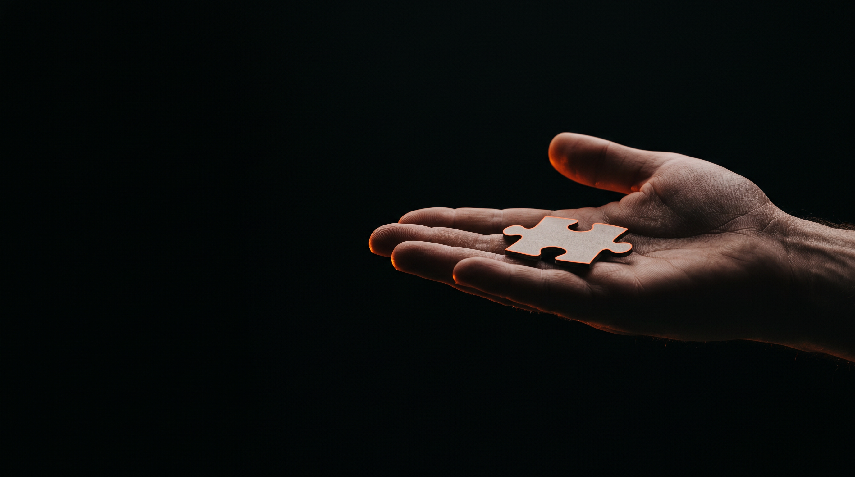 An open hand offering a single jigsaw puzzle piece forward, lit by warm orange light against deep black