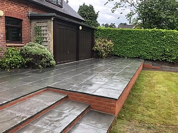 Raised dark-stone patio with brick step walls and a manicured hedge beside a brick extension with a timber door