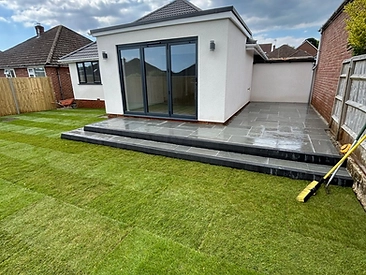 Newly landscaped rear garden with a white-rendered single-storey extension, grey stone patio and freshly laid striped lawn under a blue sky