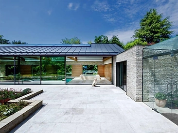 Modern home with stone-clad facade and floor-to-ceiling glass doors opening onto a light stone patio with lawn and planting
