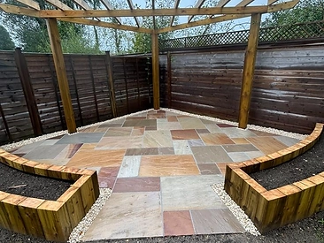 Corner timber pergola over a multi-toned stone patio, framed by raised sleeper planters and a gravel border against dark fencing