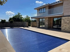 Beige stone pavers surrounding a rectangular pool with blue safety cover and modern house