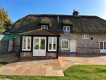 Thatched flint-and-brick cottage with a newly laid mixed-stone patio in earthy tones beside a green lawn