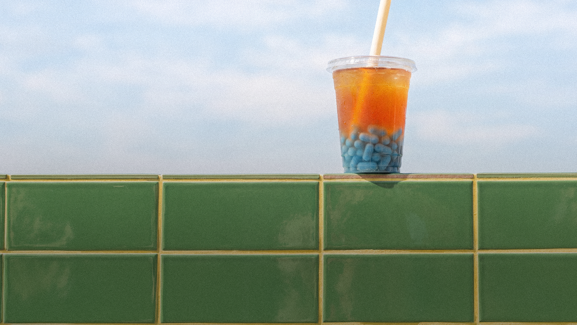 A plastic cup of bubble tea with blue PrEP pill pearls on a tiled ledge under a bright sky
