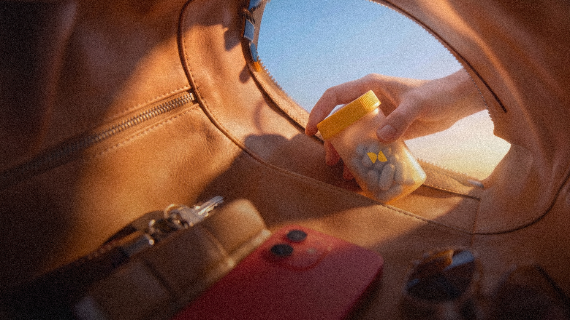 A hand holding a pill bottle inside a leather bag, open to a warm sunset sky