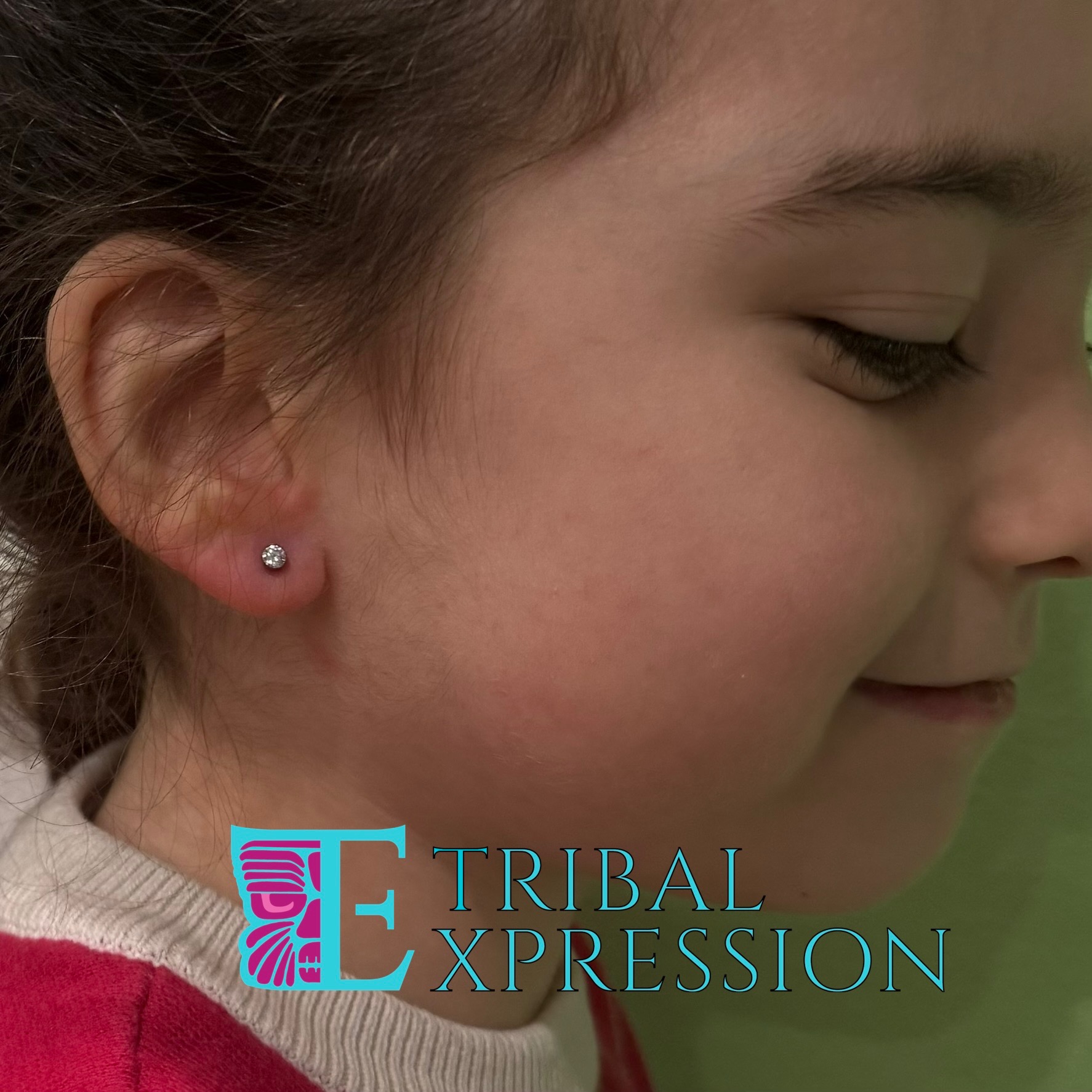 A smiling child with their eyes closed, showing off a sparkling new stud earring — a proud first-piercing moment at Tribal Expression.