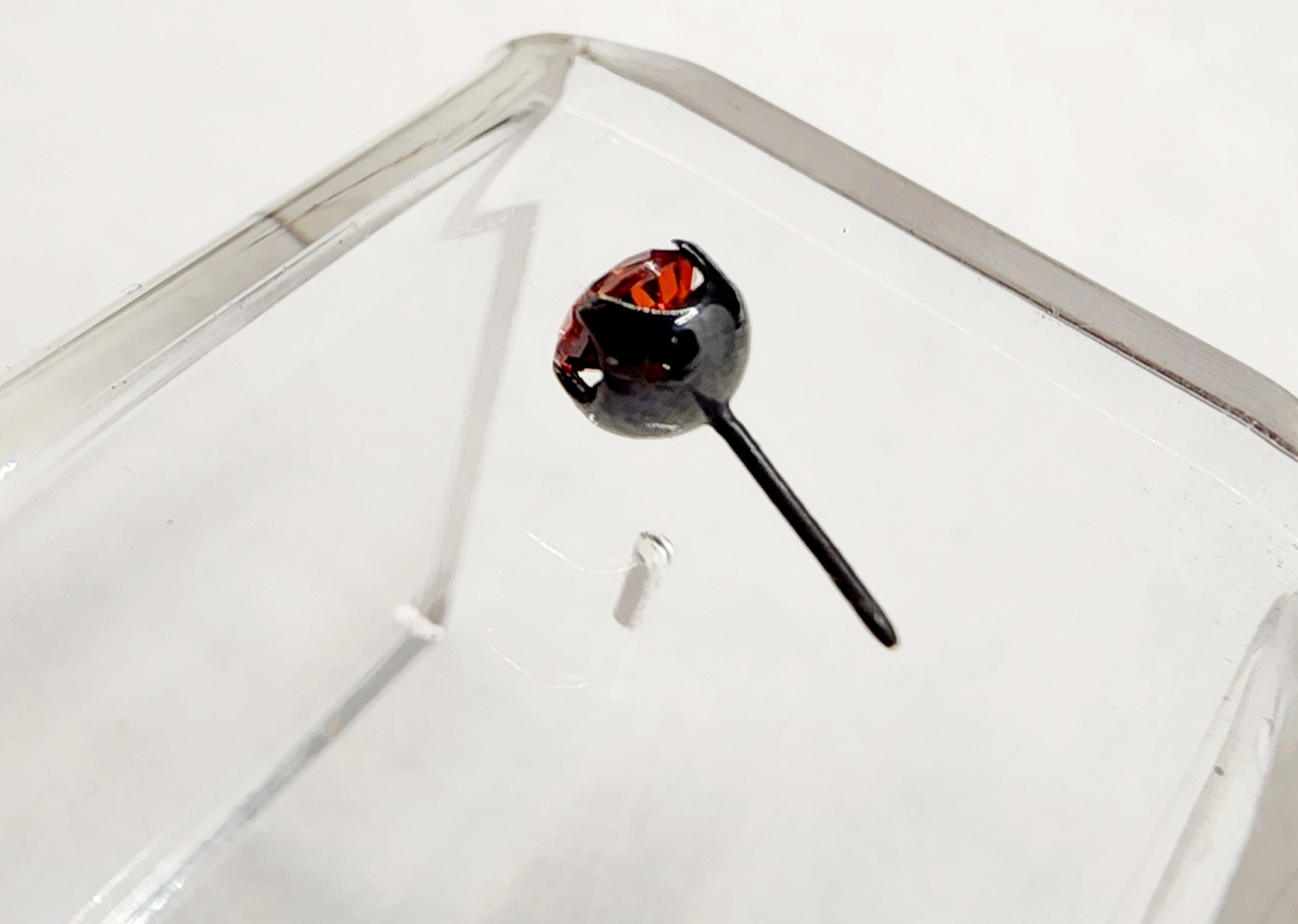 Red gemstone stud in a dark titanium setting inside a clear display case, Calgary made body jewelry