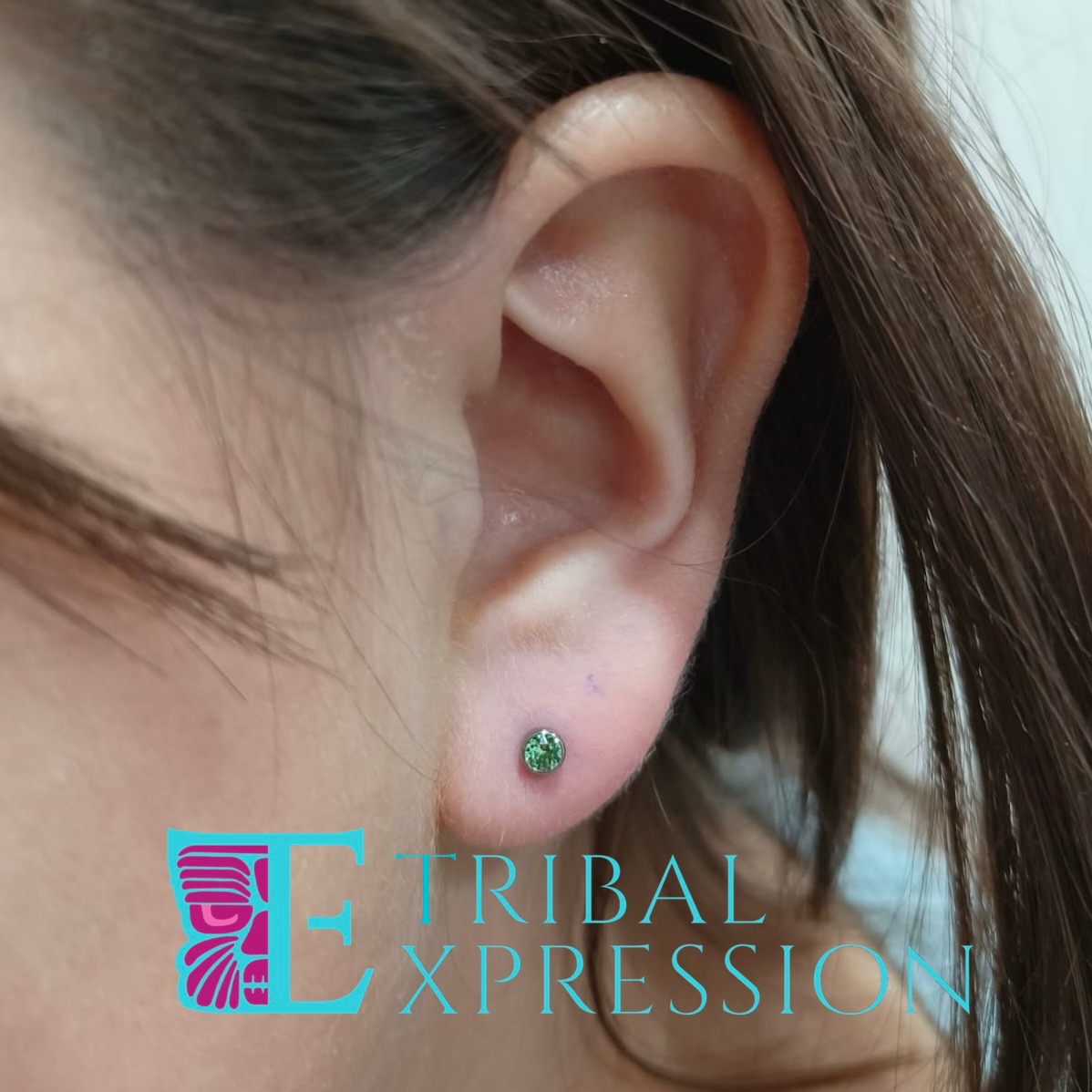 A child's ear with a green prong-set gemstone stud earring in the earlobe.