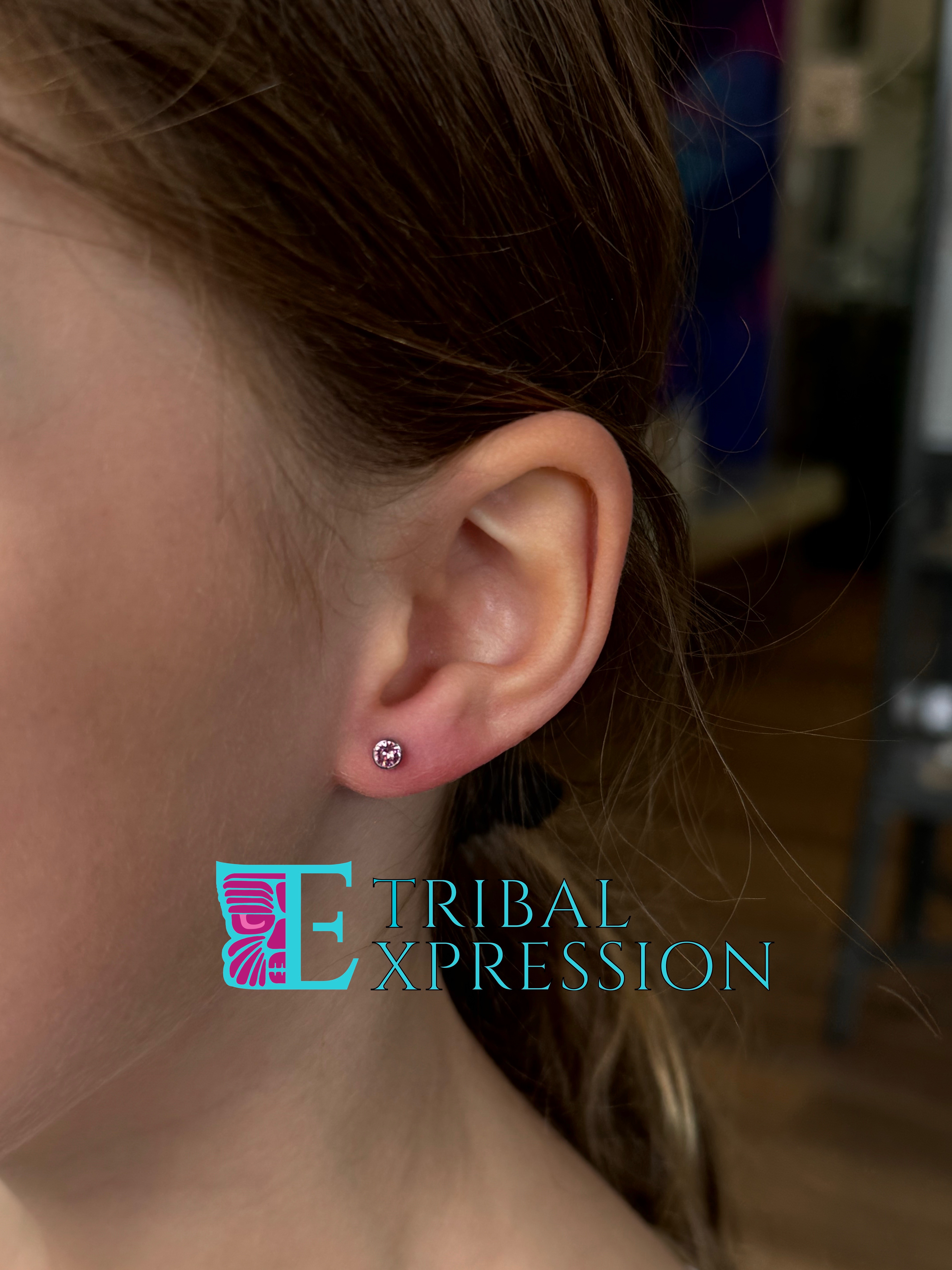A child's ear with a round pink stud earring freshly placed in the earlobe.