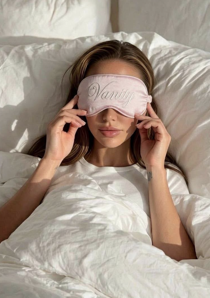 Karmel wearing a blush silk sleep mask embroidered with 'Vanity' against white bedding