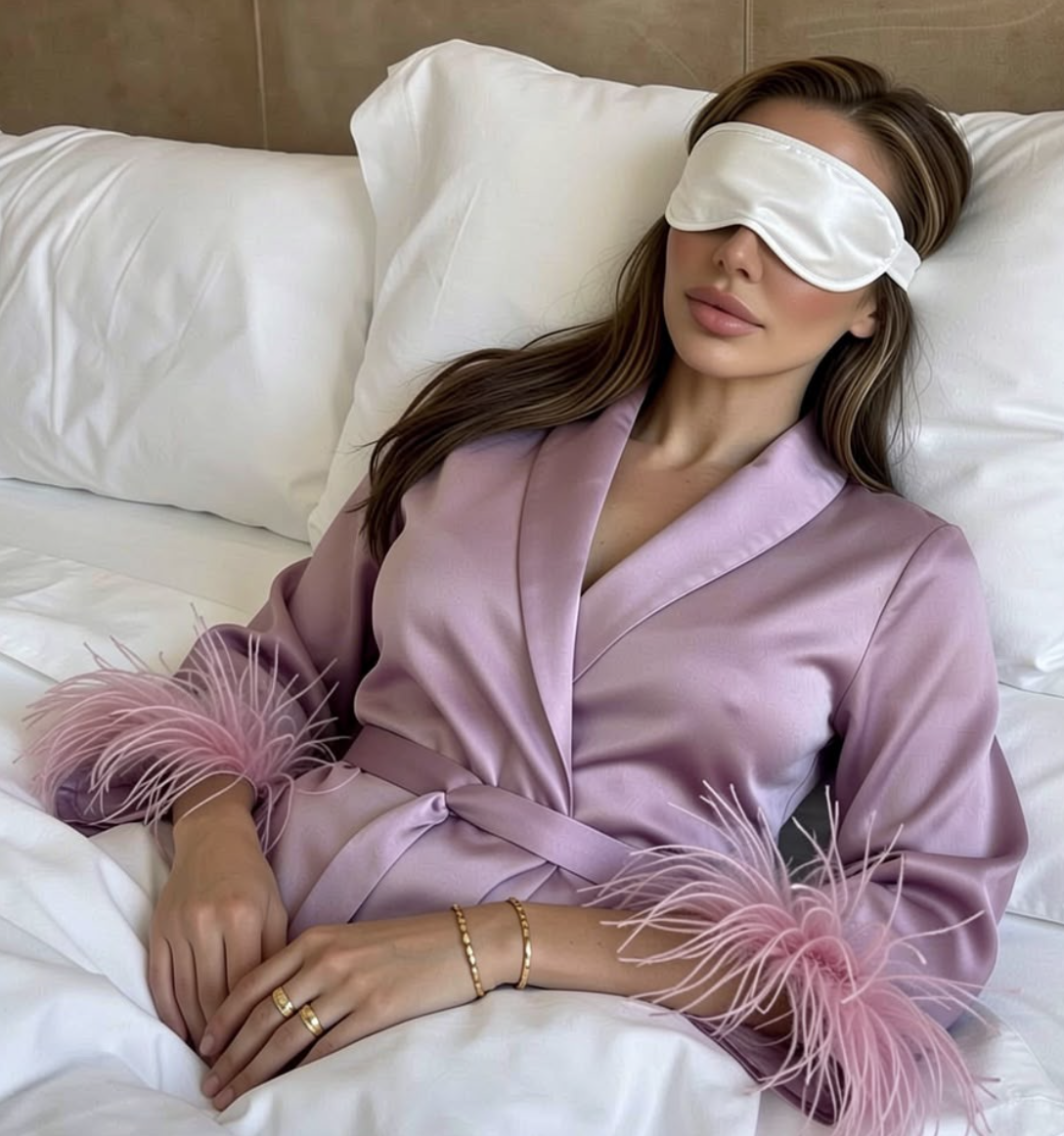 Karmel lying down in a lilac silk robe with feather trim, wearing a white sleep mask