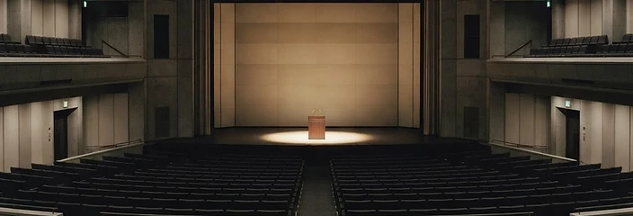 Empty auditorium stage with a single lit lectern under warm spotlight
