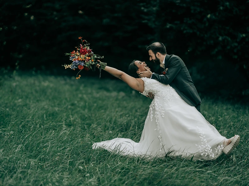 Bräutigam lässt die Braut im hohen Gras elegant zurückbeugen, dunkler Wald im Hintergrund