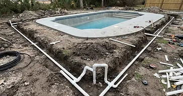 Newly excavated trench with white PVC irrigation pipes laid in it, routed around a residential swimming pool area in a rough-graded South Carolina Lowcountry backyard under construction