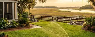 Daniel Island coastal backyard with thick green lawn, in-ground irrigation sprinkler watering grass, marsh grass and river view in the distance, live oaks