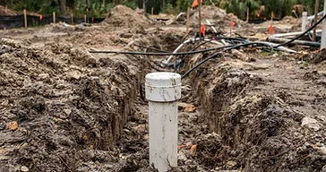 Close-up ground-level view of a white PVC irrigation pipe end with a visible PVC cap in a shallow dirt trench in a rough-graded South Carolina Lowcountry backyard under construction before pool installation