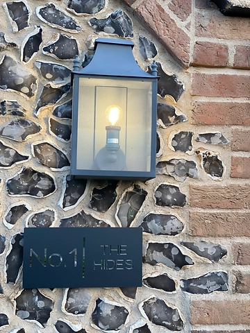 Black metal No.1 The Hides sign mounted on a flint wall beneath a lit lantern