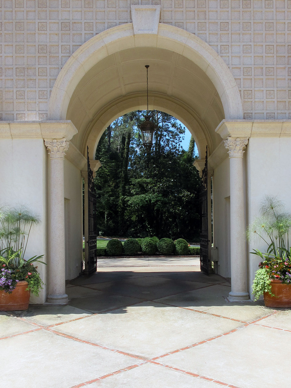 Arched stone passage with open iron gates, columns and planted pots framing a view to trees and clipped shrubs