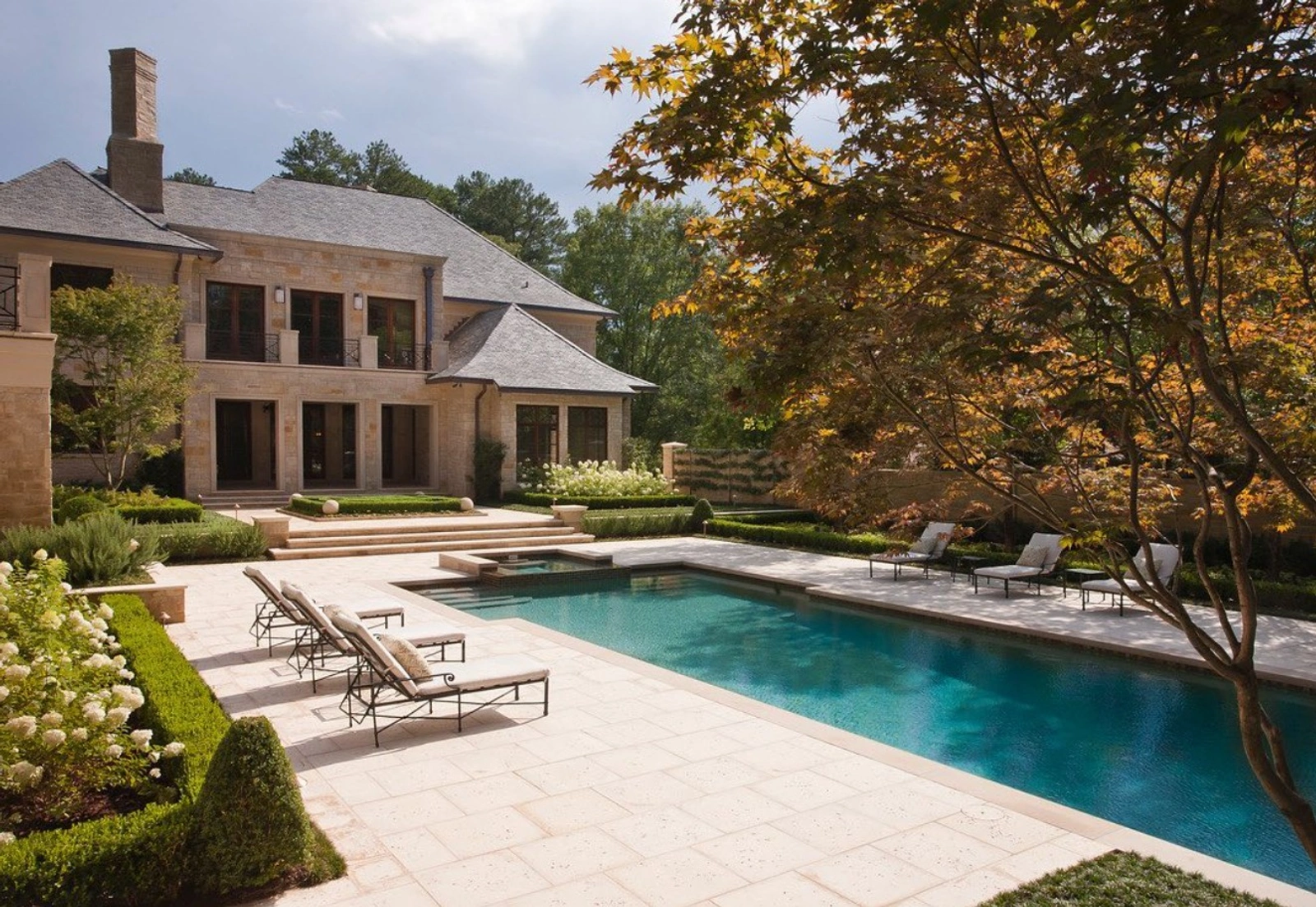 Elevated view of a stone residence with a rectangular swimming pool, lounge chairs, a boxwood hedge garden, and a mature Japanese maple tree