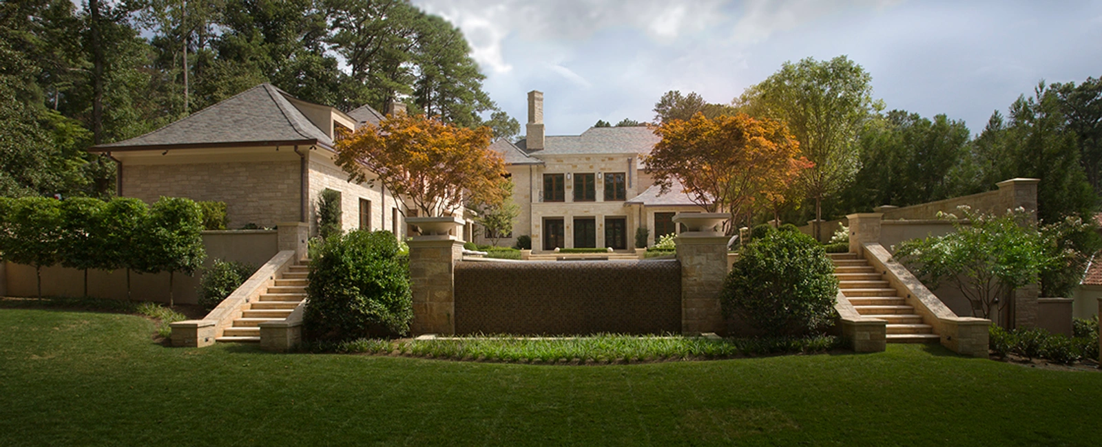 Wide view of a stone mansion with a tiered wall fountain flanked by symmetrical stone staircases, set in a wooded lot