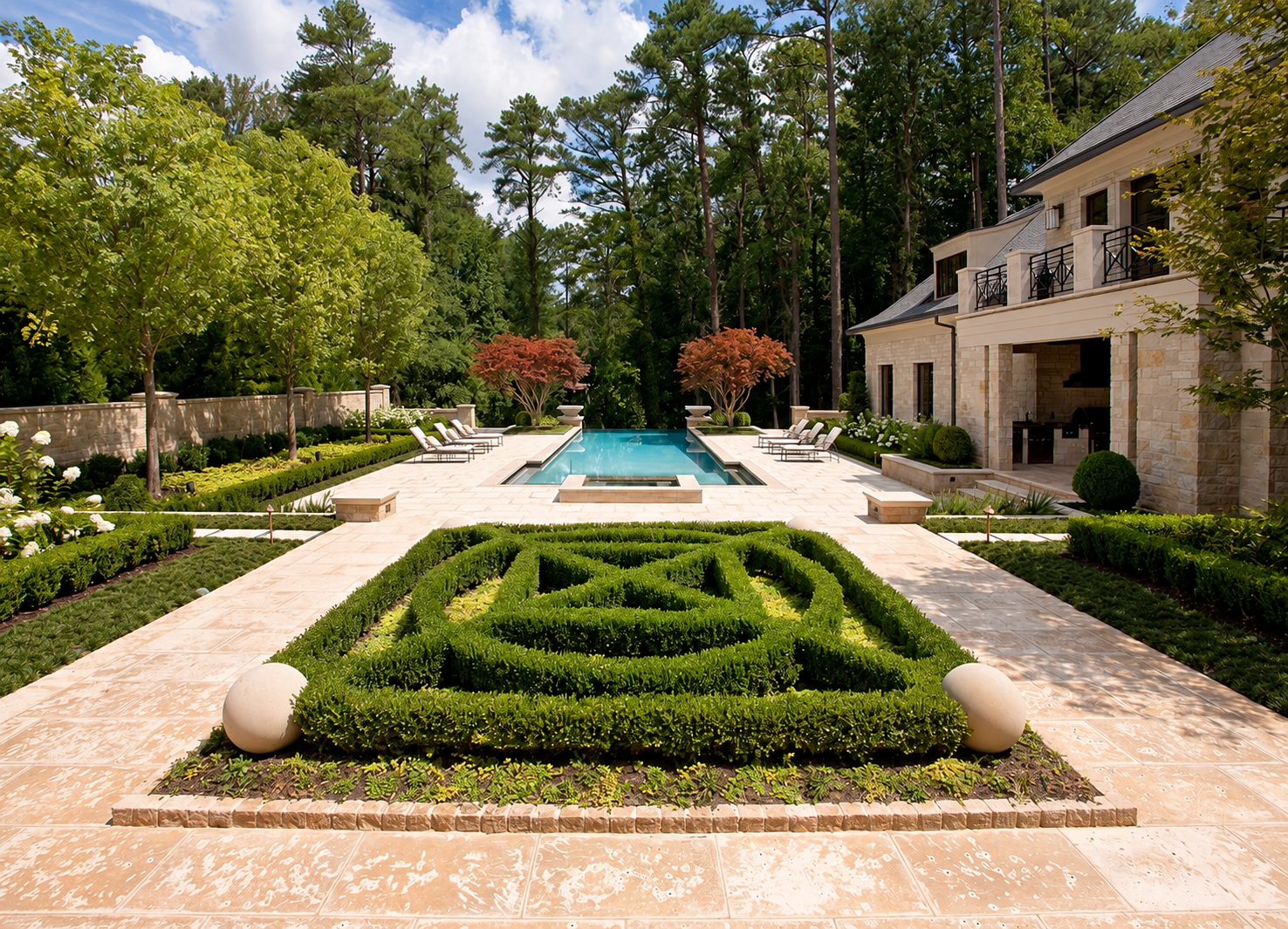 Knot garden with clipped boxwood, stone orbs, and a rectangular swimming pool bordered by travertine pavers, framed by manicured landscaping and pine forest with a stone residence beyond