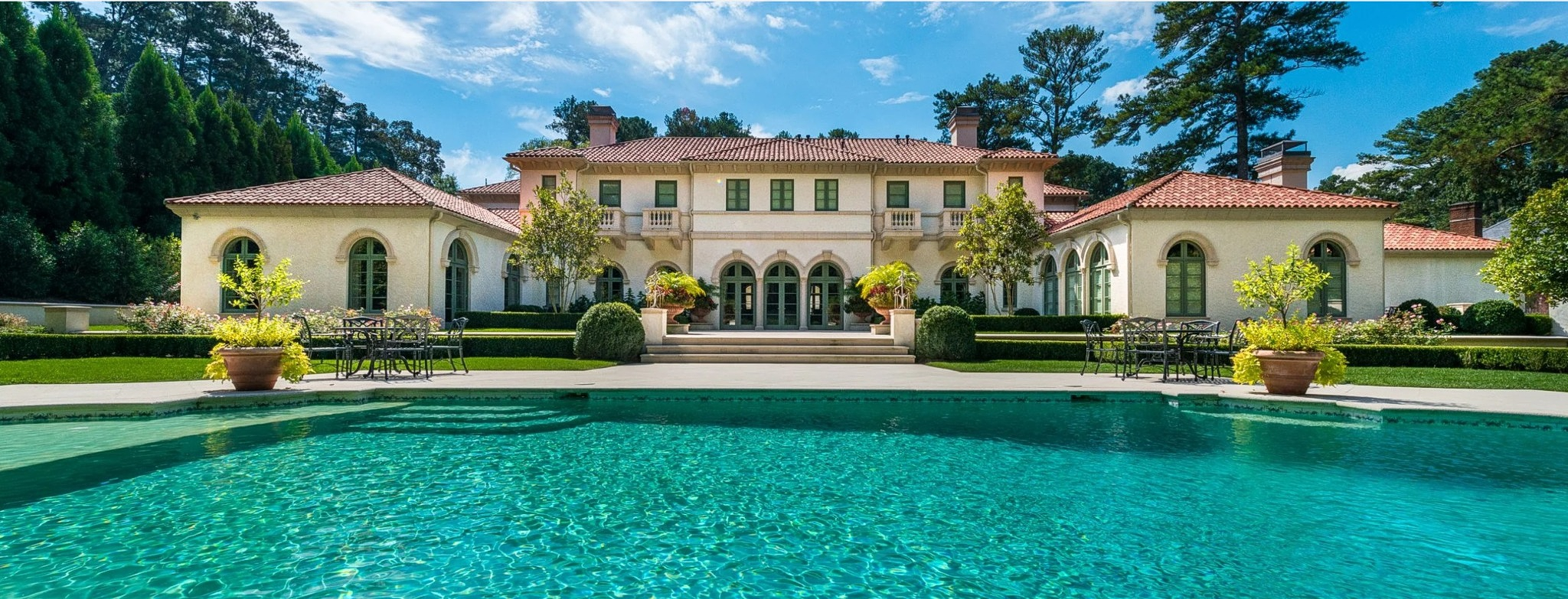 Italianate residence with tile roofs seen across a turquoise swimming pool and terrace