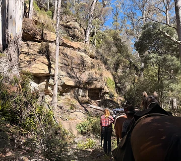 Riders on horseback following a bush trail beneath a sandstone cliff