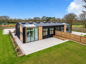 Aerial view of a modern barn-style house with a black metal standing-seam roof, light wood cladding and floor-to-ceiling glass sliding doors opening onto a paved patio