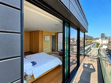 Contemporary bedroom opening through floor-to-ceiling black-frame sliding glass doors onto a weathered grey rooftop deck with a glass balustrade