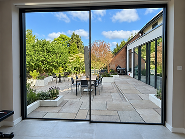 Interior of a minimalist room looking out through floor-to-ceiling minimal-frame sliding glass doors onto a sunny stone-paved garden patio
