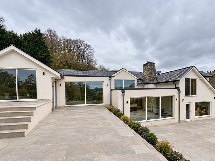 Detached house with cream rendered walls and black-framed sliding glass doors across the ground floor, opening onto a wide stone terrace with concrete steps