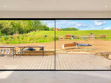 Interior of a warm-white room looking out through a two-panel black-framed sliding glass door onto a wooden deck and a green hillside beyond