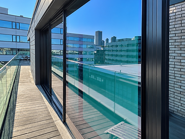 Rooftop balcony with a large black-framed floor-to-ceiling sliding glass wall reflecting the city skyline, glass balustrade and timber decking