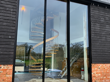 Three-panel floor-to-ceiling glass sliding door system set into a black horizontal-planked timber wall, with a curved wooden spiral staircase visible inside