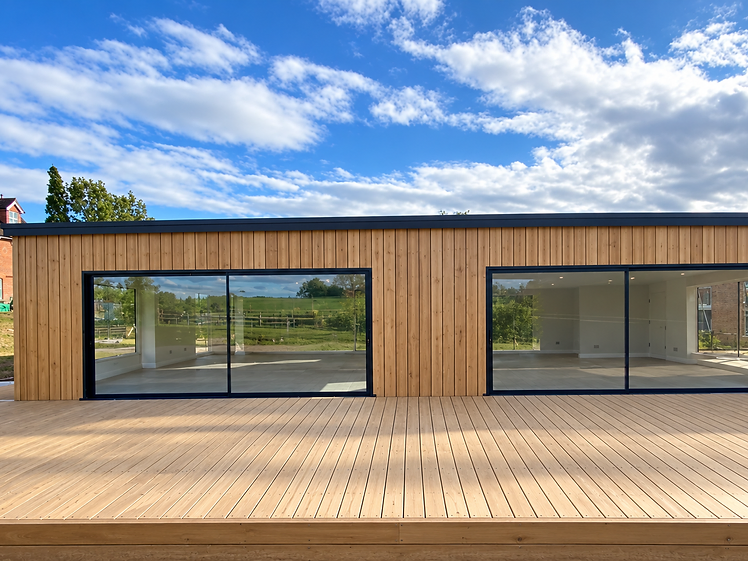 Single-storey cedar-clad extension with two floor-to-ceiling black-framed sliding glass doors opening onto a raised timber deck in bright sunlight