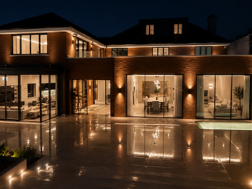 Night-time rear facade of a two-storey brick residence with extensive floor-to-ceiling minimal-frame sliding glass doors lit by exterior uplights