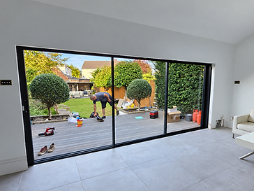 Interior of a restored room looking out through three-panel black-framed sliding glass doors onto a wooden deck with topiary trees in pots