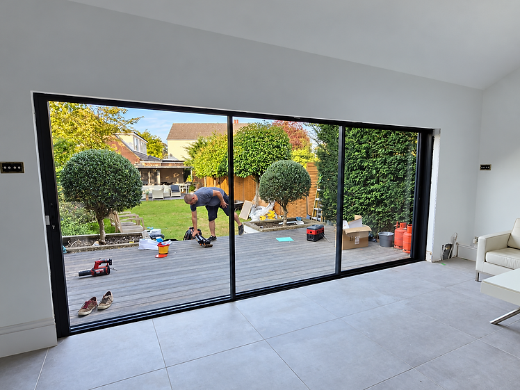 Interior of a restored room looking out through three-panel black-framed sliding glass doors onto a wooden deck with topiary trees in pots