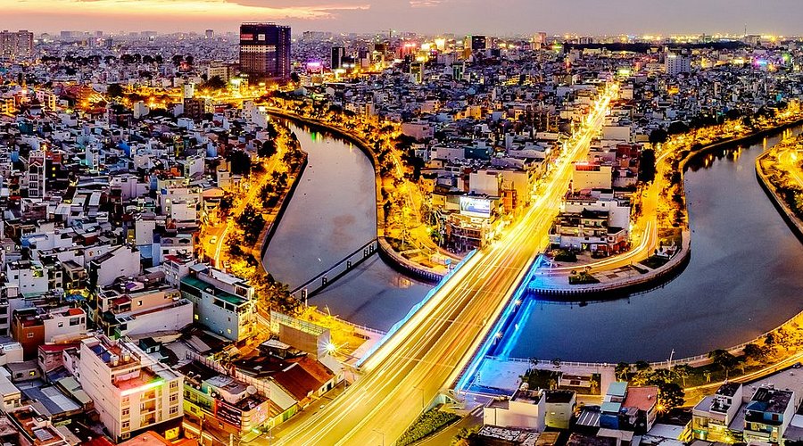 Aerial night view of a dense Vietnamese city with light trails from traffic over a bridge, representing the urban environment where digital nomads and long-term visitors rent apartments