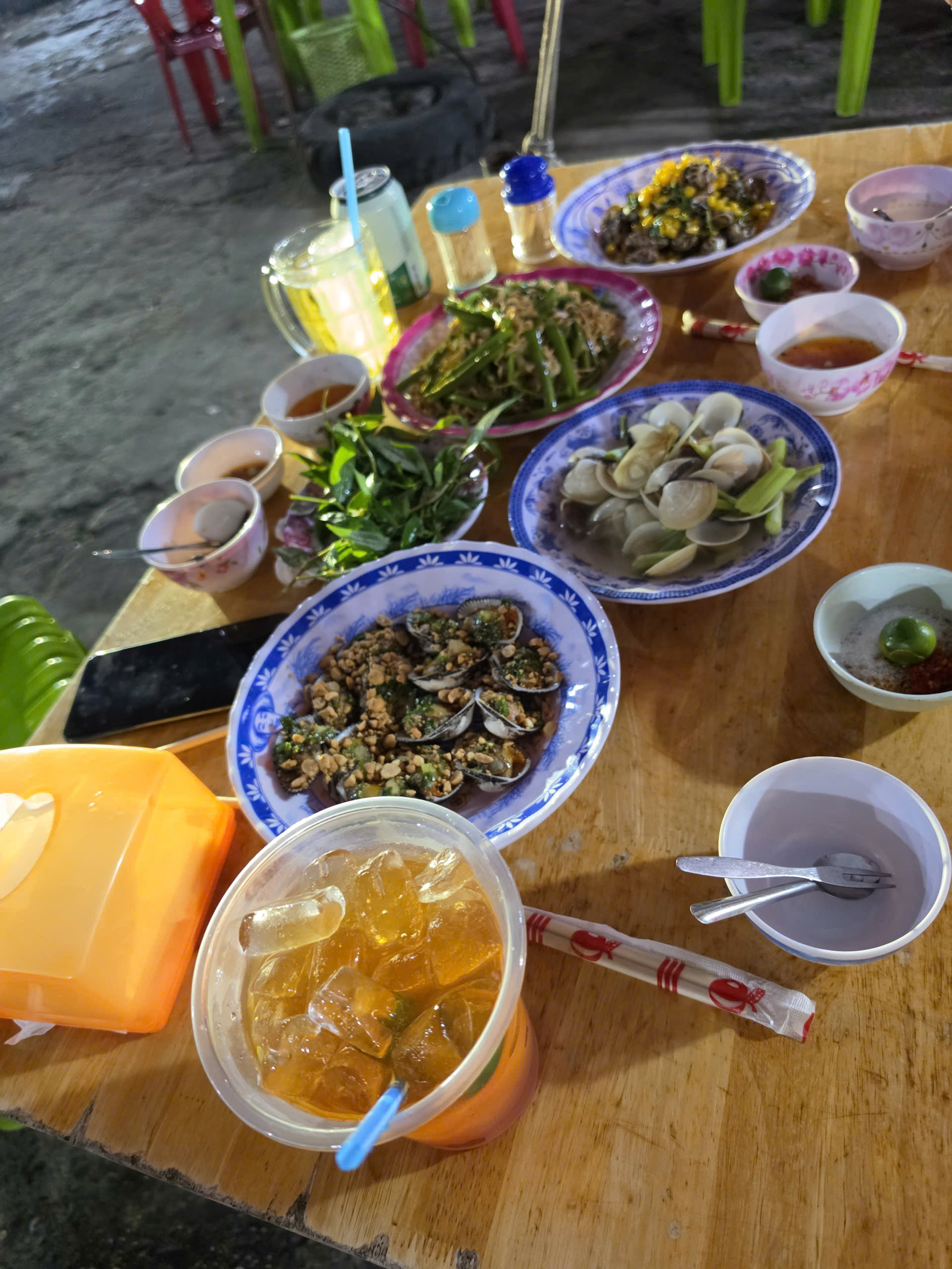 Table filled with Vietnamese seafood dishes, clams, shellfish, herbs, dipping sauces, and an iced drink