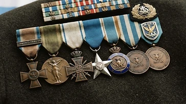 Realistic illustration of the military medals and badges awarded to Constantinos Ventiris, including Commemorative War Medal 1940–1941, Commemorative War Medal 1941–1945, Medal of Military Merit, Medal for Outstanding Acts, War Cross, Victory Medal 1914–1918, Commemorative Medal for the Greco-Bulgarian War of 1913, shown against a light Greece flag background. AI-generated illustration, not an authentic photograph.