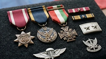 Realistic illustration of the military medals and badges awarded to Javier Rueda, including Military Medal Herido en Acción, Military Medal of Valor, Military Order of St. Matthew, shown against a light Colombia flag background. AI-generated illustration, not an authentic photograph.