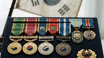 Realistic illustration of the military medals and badges awarded to Paik Sun-yup, including Order of Military Merit, Eulji, Republic of Korea Presidential Unit Citation, Republic of Korea Service Medal, Republic of Korea War Service Medal, Order of National Security Merit, Tong-Il, Order of Military Merit, Hwarang, Order of Military Merit, Chungmu, shown against a light South Korea flag background. AI-generated illustration, not an authentic photograph.