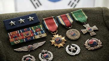 Realistic illustration of the military medals and badges awarded to Kim Hong-il, shown against a light South Korea flag background. AI-generated illustration, not an authentic photograph.