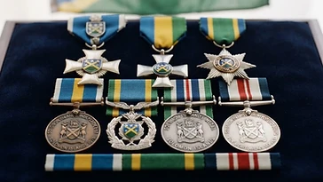 Realistic illustration of the military medals and badges awarded to Sir Peter Kenilorea, including Solomon Islands Independence Medal, Western Pacific High Commission Badge of Honour, Cross of the Solomon Islands, Star of the Solomon Islands, Solomon Islands Medal, Solomon Islands Meritorious Service Medal, shown against a light Solomon Islands flag background. AI-generated illustration, not an authentic photograph.