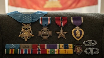 Realistic illustration of the military medals and badges awarded to Gary Michael Rose, including Medal of Honor, Distinguished Service Cross, Bronze Star Medal, Purple Heart, shown against a light Laos flag background. AI-generated illustration, not an authentic photograph.