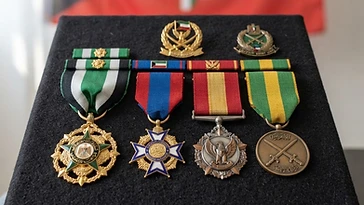 Realistic illustration of the military medals and badges awarded to Sheikh Nawaf Al-Ahmad Al-Jaber Al-Sabah, including Order of Kuwait, Order of Mubarak the Great, Order of National Defence, Order of Military Duty, shown against a light Kuwait flag background. AI-generated illustration, not an authentic photograph.